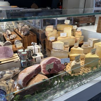 Nouvelle Maison - A glass display case showcases an assortment of cheeses, cured meats, and jars, with wooden stands and labels in a cheese shop setting. - Grocery Near Me - Winchester, Massachusetts