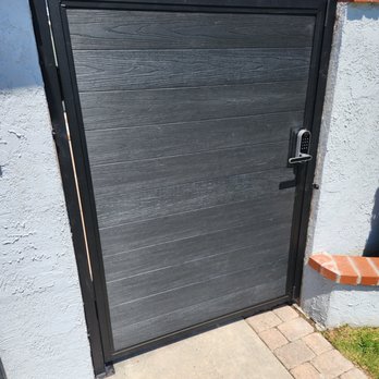 Socal Gates - A black gate with a wood grain texture features an electronic keypad lock, set between two white stucco walls, with a brick and concrete pathway in front. - Fences & Gates Near Me - Orange, California