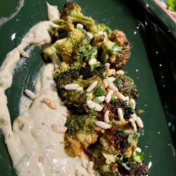 Lynette - A dish featuring roasted broccoli florets garnished with sunflower seeds and chopped herbs, served with a creamy white sauce on a green plate. The broccoli appears to be seasoned and roasted, with some parts slightly charred, and the sauce is spread alongside the vegetables, adding a contrasting texture and flavor. - Beer Bar Near Me - Minneapolis, Minnesota