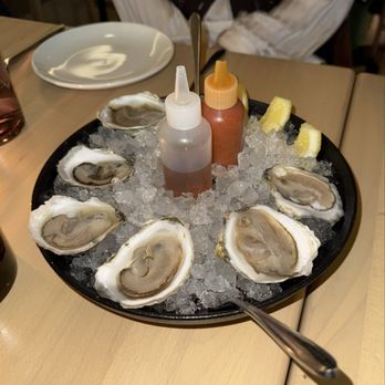 Baleia - Oysters - A platter of fresh oysters on ice with lemon wedges and condiment bottles, set on a restaurant table. - Wine Bars Near Me - Boston, Massachusetts