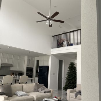 Lion Group Electrical - Removing ceiling fan and hanging 120 lb crystal chandelier - The image shows a living room with a high ceiling and a ceiling fan with dark blades. A Christmas tree is positioned near a doorway, and a beige sofa with pillows is in the foreground. The room opens into a kitchen area with white cabinetry, a black refrigerator, and a breakfast bar with chairs. An upper-level balcony with a black railing is visible, with some items stored on it. The space is well-lit with natural light. - Electricians Near Me