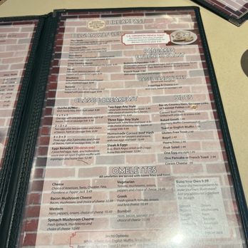 Sunset Grill - The menu lists breakfast options such as Belgian waffles, pancakes, French toast, bagel sandwiches, and omelettes. It provides detailed descriptions and prices for each item, including specialty options like the Bomber omelette and Blueberry pancakes. The menu is printed on a paper sheet inside a plastic cover, with a brick wall background visible in the image. - American Near Me - Ithaca, New York
