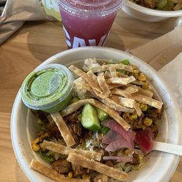 Cava Bowl - A food bowl with grilled meat, vegetables, and crispy strips, served with a green sauce, alongside a pink beverage in a plastic cup.