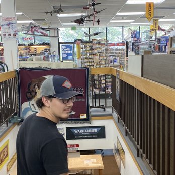 HobbyTown - A man with glasses, a black cap, and a black T-shirt stands in an aisle of a hobby store, facing slightly to the left. The store has model airplanes hanging from the ceiling and shelves stocked with hobby supplies, including model kits and accessories. Large windows at the back let in natural light, and signs indicating store policies and product categories are visible. The store appears organized with a variety of hobby-related items on display. - Toy Stores Near Me - Aurora, Colorado