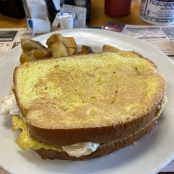 Bea's Cafe - A breakfast meal on a white plate includes a fried egg sandwich with thick toasted bread, fried eggs, and possibly bacon or sausage. Accompanying the sandwich are fried potato chunks. The table has a paper menu, a red cup, a salt shaker, and a drink container in the background, indicating a casual dining setting. - Breakfast & Brunch Near Me - Conway, New Hampshire