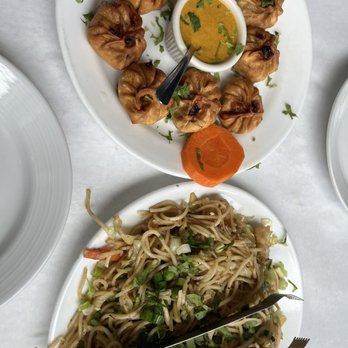 Nepal House - The image shows a white plate of fried momos arranged in a circle around a small bowl of dipping sauce, garnished with cilantro and a slice of carrot. Below, there is another white plate filled with stir-fried noodles mixed with vegetables and chopped green onions. The dishes are placed on a white tablecloth, indicating a meal at a restaurant offering Asian cuisine. - Indian Near Me - Chicago, Illinois