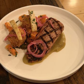 Linden + Front - Pork tenderloin with smoked potato croquettes - A white plate holds grilled meat with grill marks, pickled red onions, and roasted vegetables, served on a wooden table. - New American Near Me - Bath, Maine
