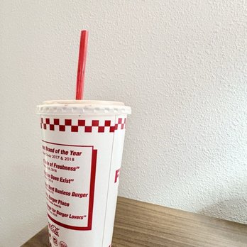 Five Guys - Chocolate shake!! Yum! - A large soda cup with a red straw is positioned on a wooden table. The cup has a white background with red checkered patterns and text, and it is set against a plain, textured white wall. The cup appears to be from a fast-food restaurant, and the image is taken indoors with soft lighting. - Burgers Near Me - Malta, New York