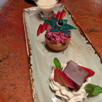 Desert Bistro - A rectangular ceramic plate holds three desserts: a berry-topped cupcake with pink frosting and decorative chocolate, a chocolate and strawberry dessert with whipped cream, and a small glass of panna cotta with sliced strawberries. - New American Near Me - Moab, Utah