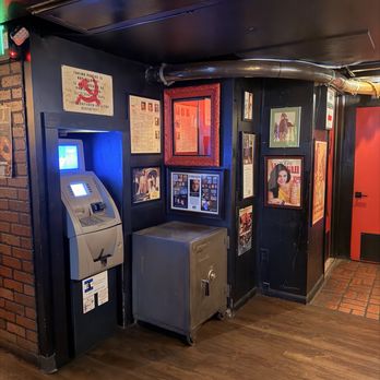 SafeHouse - The image depicts an ATM machine and a large, silver safe placed against a black wall. The wall is decorated with framed posters, photographs, and newspaper clippings, creating a themed ambiance. The floor is wooden, and there is a brick wall visible on the left side. The setting appears to be inside a bar or restaurant with a spy or entertainment theme. - Burgers Near Me - Milwaukee, Wisconsin