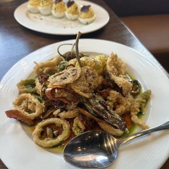 Entourage - Calamari - A white plate contains fried calamari rings mixed with sliced peppers and herbs, served with a sauce or oil. In the background, a white oval dish holds deviled eggs topped with garnishes. The setting appears to be a restaurant table with a dark wooden surface, and part of a person’s arm and clothing are visible. - American Near Me - Downers Grove, Illinois