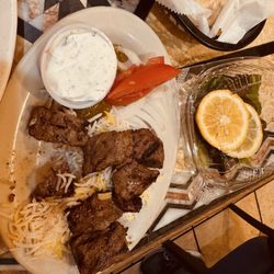 Fadi's Meyerland Mediterranean Grill - Need Beef Kabob (Per Skewer)en una cama de arroz con rebanadas de cebolla y tomate y pepinillo, y salsa tatziki