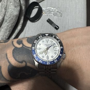 Watch Outlet - A Seiko automatic GMT wristwatch with a white dial, black and blue bezel, and metal bracelet is worn on a person's wrist with a tattoo. - Watches Near Me - Patchogue, New York