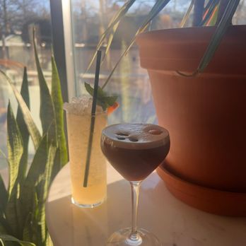 The Snail - Carajillo Martini and cocktail - A dark cocktail in a stemmed glass and a light-colored cocktail in a tall glass are positioned on a white table near a window. The sunlight streams through the window, illuminating the drinks and casting shadows. Potted plants are visible, with one large plant partially obscuring the view outside, which includes trees and a clear sky. The scene suggests a relaxed setting, possibly in a restaurant or cafe. - American Near Me - Brooklyn, New York