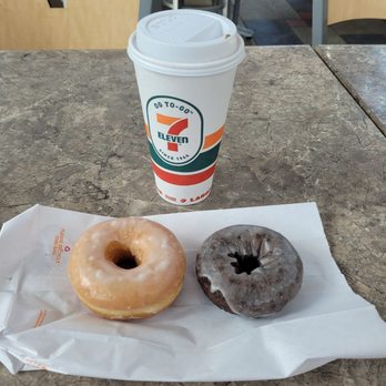 Dunkin' - Winning combo: 7-Eleven Coffee and Dunkin' Dodos - A paper wrapper on a table holds two donuts, one glazed and one chocolate, alongside a large coffee cup from 7-Eleven. The donuts are positioned side by side, with the glazed donut on the left and the chocolate donut on the right. The coffee cup features the 7-Eleven logo and is topped with a plastic lid. The background shows chairs and a tiled floor, indicating a casual dining or coffee area. - Donuts Near Me - Waterfall, Pennsylvania