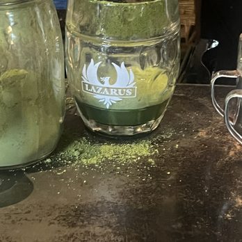 Lazarus Brewing - A glass with the Lazarus Brewing logo contains a dark liquid and green powder, with some powder spilled on the wooden table surface. - Breweries Near Me - Austin, Texas