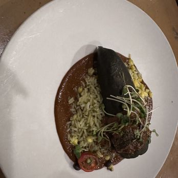 Amaya - Poblano - A plated Mexican dish includes a large poblano pepper stuffed with filling, placed on a bed of red sauce. The plate also contains rice mixed with corn and garnished with microgreens and tomato slices. The presentation is on a white, oval-shaped plate, with a brown table surface visible in the background. - New American Near Me - Santa Fe, New Mexico