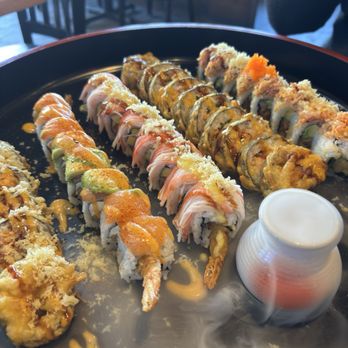Koi Sushi - Dragon Roll(8pcs) Bears Roll(6 pcs) Godzilla Roll(10pcs) Sweet Heart Roll(10pcs) - A large black tray holds various sushi rolls, including tempura, seafood, and vegetable options, with a small container of dipping sauce. - Japanese Near Me - Chicago, Illinois