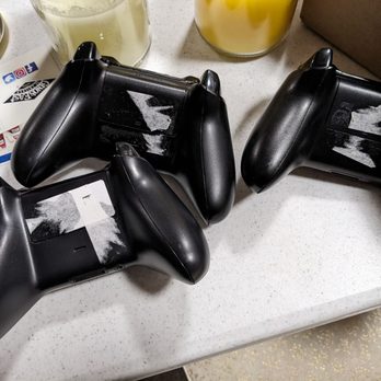 uBreakiFix by Asurion - The battery compartment on the left one is not even accessible, the label has to be peeled further. - Three black gaming controllers with tape on their screens are placed on a white surface, surrounded by drinks and miscellaneous items. - Electronics Repair Near Me - Matteson, Illinois