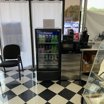 Shopper's Kitchen - Covidlly correct drinks...I love it - A glass-front beverage refrigerator containing various soda bottles and cans is situated on a checkered black and white floor. To the left, there is a black chair with armrests. To the right, a counter holds coffee-making equipment, including a coffee machine and other supplies. A window behind the refrigerator shows parked cars outside, and a sign on the window lists prices for water, apple juice, soda, and lemonade/tea. - Desserts Near Me - Topeka, Kansas