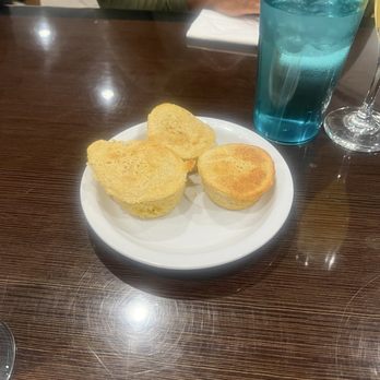 Cajun Breakfast Joint - Complimentary Praline Cornbread - Three pieces of cornbread are served on a white plate, accompanied by a tall blue drink and a wine glass, on a dark wooden table. - Breakfast & Brunch Near Me - Phoenix, Arizona