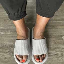 Beautiful Nails! Yes, freshly done at Spring Ridge Nails! - The image shows a person's feet in white textured slides with open toes, revealing toenails painted bright orange. The person is wearing black pants with elastic cuffs at the ankles. The background features a wooden floor with a natural grain pattern. The photo is taken from a top-down perspective, focusing on the footwear and feet.