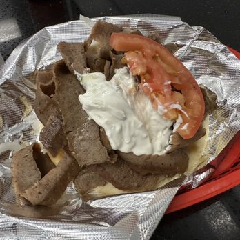George's Gyros - a plate of food - A portion of sliced gyro meat topped with a slice of tomato, onion, and a dollop of white sauce, served on foil-lined paper inside a red plastic basket. The background features a dark countertop, indicating a casual dining or takeout setting at a gyro restaurant. - American Near Me - Chicago, Illinois