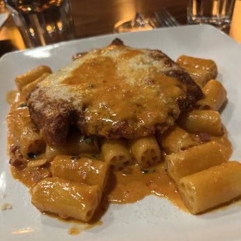 Carmine - Chicken vodka sauce - A dish featuring rigatoni pasta coated in a creamy, orange-colored sauce, topped with a breaded chicken cutlet with melted cheese. The plate is set on a table with glasses of water and wine in the background, indicating a dining environment. - Italian Near Me - Newburyport, Massachusetts