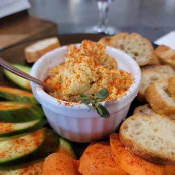 Mindscape Fermentations - A food presentation includes a small white ramekin of hummus sprinkled with paprika, surrounded by sliced cucumbers, bread slices, and crackers on a plate. A glass of red wine is visible in the background, placed on a table with a blurred setting. - Breweries Near Me - Rocklin, California