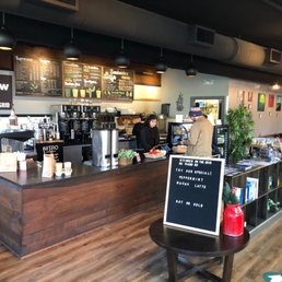 Order and serving area - Coffee shop interior featuring a wooden counter with baristas preparing drinks, menu boards above listing coffee options, and customers placing orders. A small table with a sign promoting special drinks and a red container with holiday decorations is in the foreground. The space includes seating areas, artwork on the walls, and a cozy atmosphere.
