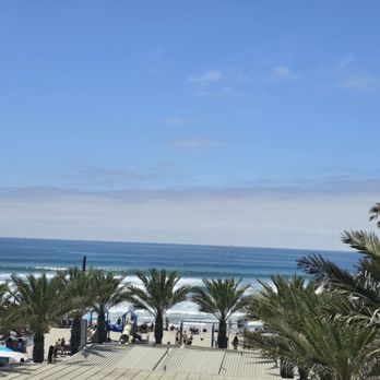 High/Low - View - The image shows a beachfront view with multiple palm trees along the shoreline, ocean waves rolling onto the sand, and a partly cloudy blue sky. The photo appears to be taken from an elevated vantage point, possibly from a balcony or rooftop, overlooking the beach area with people and structures visible near the shore. - Breakfast & Brunch Near Me - Oceanside, California