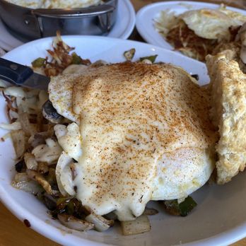The Southern Cafe - Stuffed Cajun Cheese Steak Biscuit - A breakfast dish includes a fried egg seasoned with paprika, placed over sautéed vegetables such as mushrooms, onions, and green peppers. The dish is served in a white bowl, with a biscuit on the side. In the background, there are additional breakfast items, including a skillet with eggs and a plate with fried eggs and other breakfast foods. The setting appears to be a casual restaurant or cafe. - Southern Near Me - Roselle, Illinois