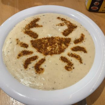 Koco's Pub - Creamy Crab soup - A white bowl contains creamy crab soup with a smooth texture. The surface of the soup has a face design made with brown spices, including eyes, nose, and mouth. The bowl is placed on a wooden table, with some food packaging visible in the background. - Pubs Near Me - Baltimore, Maryland