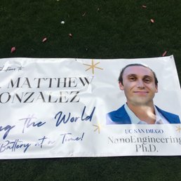 A printed banner celebrating Dr. Matthew Gonzalez, a UC San Diego NanoEngineering Ph.D., is laid on grass with scattered flower petals.