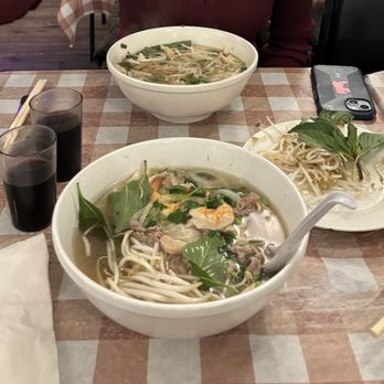 Tram's Kitchen - Two bowls of Vietnamese pho with fresh herbs and bean sprouts, along with two glasses of dark soda, are on a checkered table at a restaurant. - Vietnamese Near Me - Pittsburgh, Pennsylvania