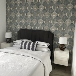 Wallpaper and paint transformed this bedroom - thanks JT Express Solution! - The image shows a bedroom with a black, vertically paneled upholstered headboard against a wall decorated with geometric-patterned wallpaper in shades of gray and black. On each side of the bed, there are white nightstands with brown, geometric-shaped lamps and white lampshades. The bed is made with white bedding and a decorative black-and-white pillow. The floor has light-colored wood or laminate flooring, and a small beige rug is partially visible at the foot of the bed.