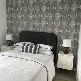 JT Express Solutions - Wallpaper and paint transformed this bedroom - thanks JT Express Solution! - The image shows a bedroom with a black, vertically paneled upholstered headboard against a wall decorated with geometric-patterned wallpaper in shades of gray and black. On each side of the bed, there are white nightstands with brown, geometric-shaped lamps and white lampshades. The bed is made with white bedding and a decorative black-and-white pillow. The floor has light-colored wood or laminate flooring, and a small beige rug is partially visible at the foot of the bed. - Flooring Near Me
