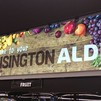 ALDI - Aldi letting everyone know it's Kensington (also known as North Bethesda) - The image shows an illuminated store sign inside ALDI in Kensington, MD, featuring the text "Welcome to your KENSINGTON ALDI" on a wooden background. The sign is decorated with images of fresh produce such as grapes, apples, peppers, and leafy greens, indicating the store's focus on groceries and fresh food offerings. - Grocery Near Me - Kensington, Maryland