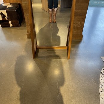 Tecovas - The image shows a person standing in front of a full-length mirror with a wooden frame, capturing their reflection. The person is wearing a green top, dark pants, and cowboy boots. The store interior includes a cowhide-patterned ottoman on the left and a wooden counter in the background. The floor is polished concrete with visible cracks, and natural light enters from a door on the right. - Leather Goods Near Me - Denver, Colorado