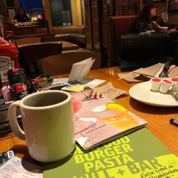 Applebee's Grill + Bar - How I like to Applebees - The image shows the interior of an Applebee's restaurant with a wooden table in the foreground, featuring menus, condiments, and a white coffee mug. In the background, a woman is seated at a table, drinking water, with other patrons and restaurant decor visible. The setting appears to be during evening hours, with warm lighting and a casual dining atmosphere. - Sports Bars Near Me - Kennett Square, Pennsylvania