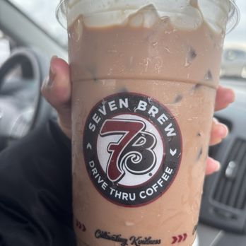 7 Brew Coffee - German chocolate drink - A person is holding a large iced coffee from Seven Brew Coffee in a clear plastic cup with a lid. The cup features the Seven Brew logo and the slogan "Drive Thru Coffee." The beverage appears to be a coffee-based drink with ice, and condensation is visible on the outside of the cup. The background shows the interior of a vehicle, including part of the dashboard and steering wheel, with a cloudy sky visible through the windshield. - Coffee & Tea Near Me - Huntley, Illinois