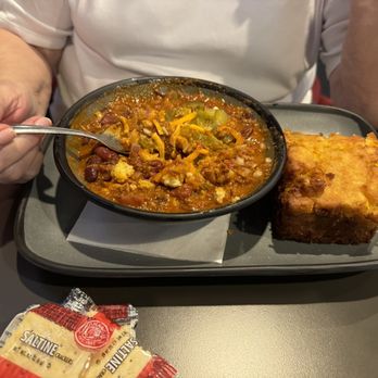 TJ Chumps - Big bowl of chili with jalapeño cornbread - A person is seated at a table, holding a spoon over a bowl of chili that contains beans, vegetables, and shredded cheese. The chili is served on a black tray with a piece of cornbread placed beside the bowl. A packet of saltine crackers is also visible on the table. - Chicken Wings Near Me - Kettering, Ohio