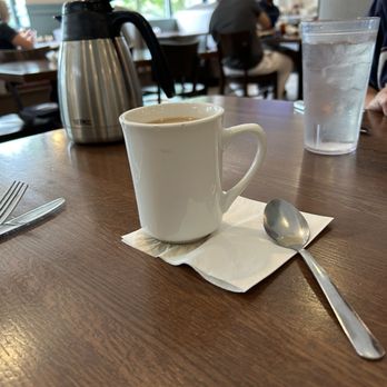 Pomegranate Restaurant - Coffee served with a backup urn - A white coffee mug, a metal spoon on a napkin, and a glass of water are on a restaurant table. A coffee pot is also visible. - American Near Me - Aurora, Illinois
