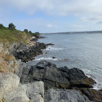 Cliff Walk - The image depicts a rugged coastline with large rocks and boulders along the shore, adjacent to the ocean. The water is calm with gentle waves, and a distant shoreline is visible across the water. The sky is partly cloudy with patches of blue, and some greenery is present on the land above the rocks. - Parks Near Me - Newport, Rhode Island