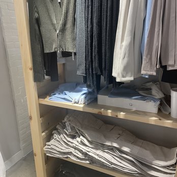 Garage - The image shows a retail clothing display with various garments. Sweaters in gray and black are hanging on hangers, while shirts and pants are folded on wooden shelves below. The display is organized with sweaters on the upper rack and pants on the lower shelves, set against a white brick wall background. - Women's Clothing Near Me - Columbus, Ohio