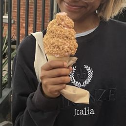 Cake Cone - A person is holding a fried chicken cone with a waffle cone base, outdoors near a brick building and metal railing. The individual is wearing a black sweatshirt with a white emblem and the word 'Italia' on it. The person's face is partially visible, showing a smile and blonde hair. The background includes brick walls and some greenery, indicating an outdoor dining or casual area.