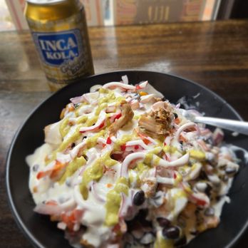 Culantro - Cilantro Bowl. Inca Cola - A black bowl contains a salad topped with creamy dressing, shredded vegetables, beans, and pieces of chicken. A yellow-green sauce is drizzled over the salad. In the background, a can of Inca Kola soda is placed on a wooden table near a window with colorful patterned curtains. - Peruvian Near Me - Ann Arbor, Michigan