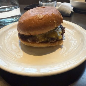 Lynette - A cheeseburger with melted cheese covering a beef patty is placed on a white ceramic plate. The burger has a toasted bun top and appears to be served in a restaurant setting, with glasses of water, a napkin, and other tableware visible in the background. - Beer Bar Near Me - Minneapolis, Minnesota