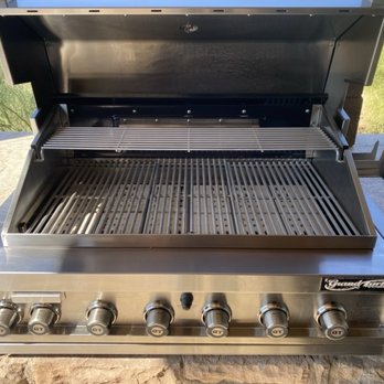 BBQ Cleaning Guru - A stainless steel gas grill featuring six control knobs labeled 'GT' and a warming rack, situated outdoors on a stone surface. The grill has a lid that is open, revealing cooking grates and a warming rack. The background includes green trees and natural scenery, indicating an outdoor setting. - Grill Services Near Me - Phoenix, Arizona