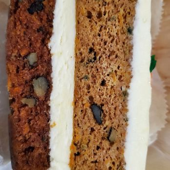 Bread & Roses - Carrot cake - A photograph shows a slice of carrot cake with cream cheese frosting layered on top. The cake contains visible nuts and dark chocolate chips, with a moist texture. It is placed on a clear, ridged glass plate, and the background appears to be a wooden surface. The image emphasizes the cake's ingredients and presentation. - Coffee & Tea Near Me - Wells, Maine